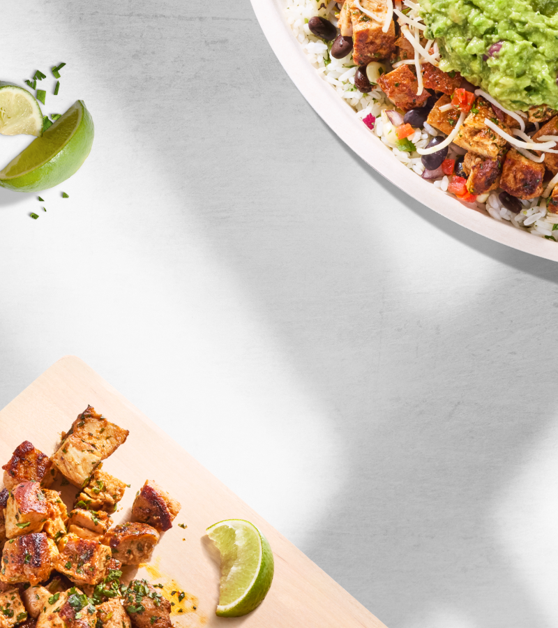 A Chipotle Pollo Asado bowl resting on a table next to a cutting board with freshly made Pollo Asado and a slice of lime.